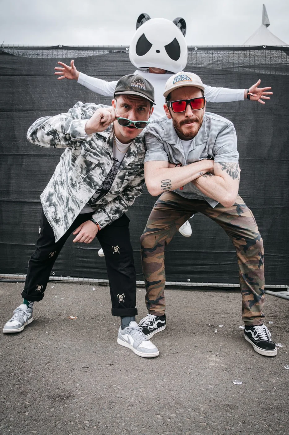 Two musicians in sunglasses striking a pose backstage at a festival, with a person in a giant panda mask spreading their arms behind them